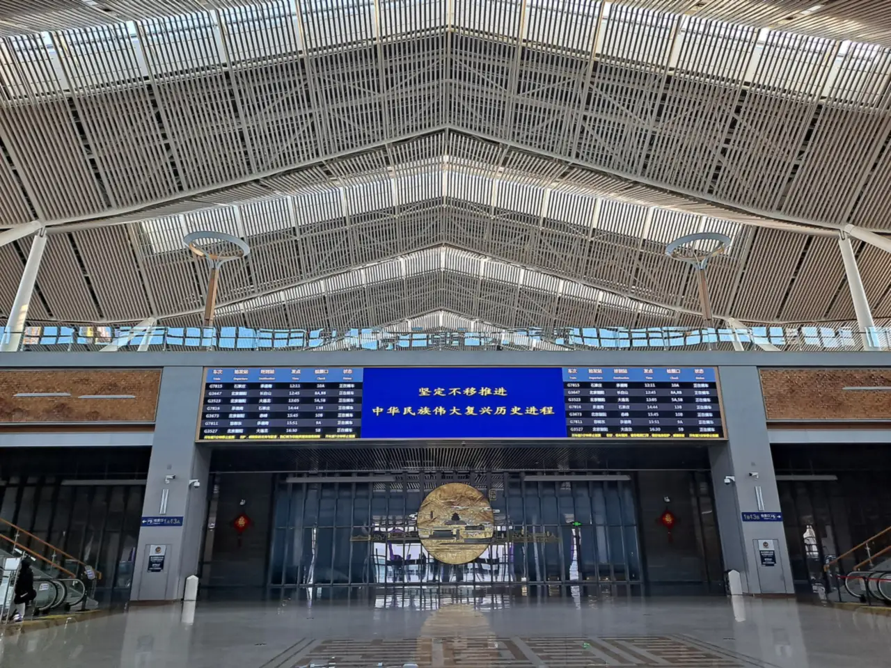 The departure hall of a railway station with a lattice roof, a large departures board and a round bronze relief above the doors