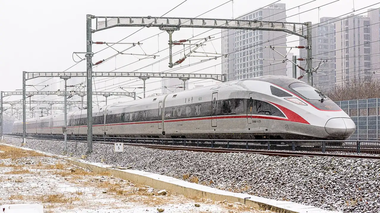 A silver and red Fuxing high-speed train running past snow-dusted ballast and bare grass with apartment blocks behind it