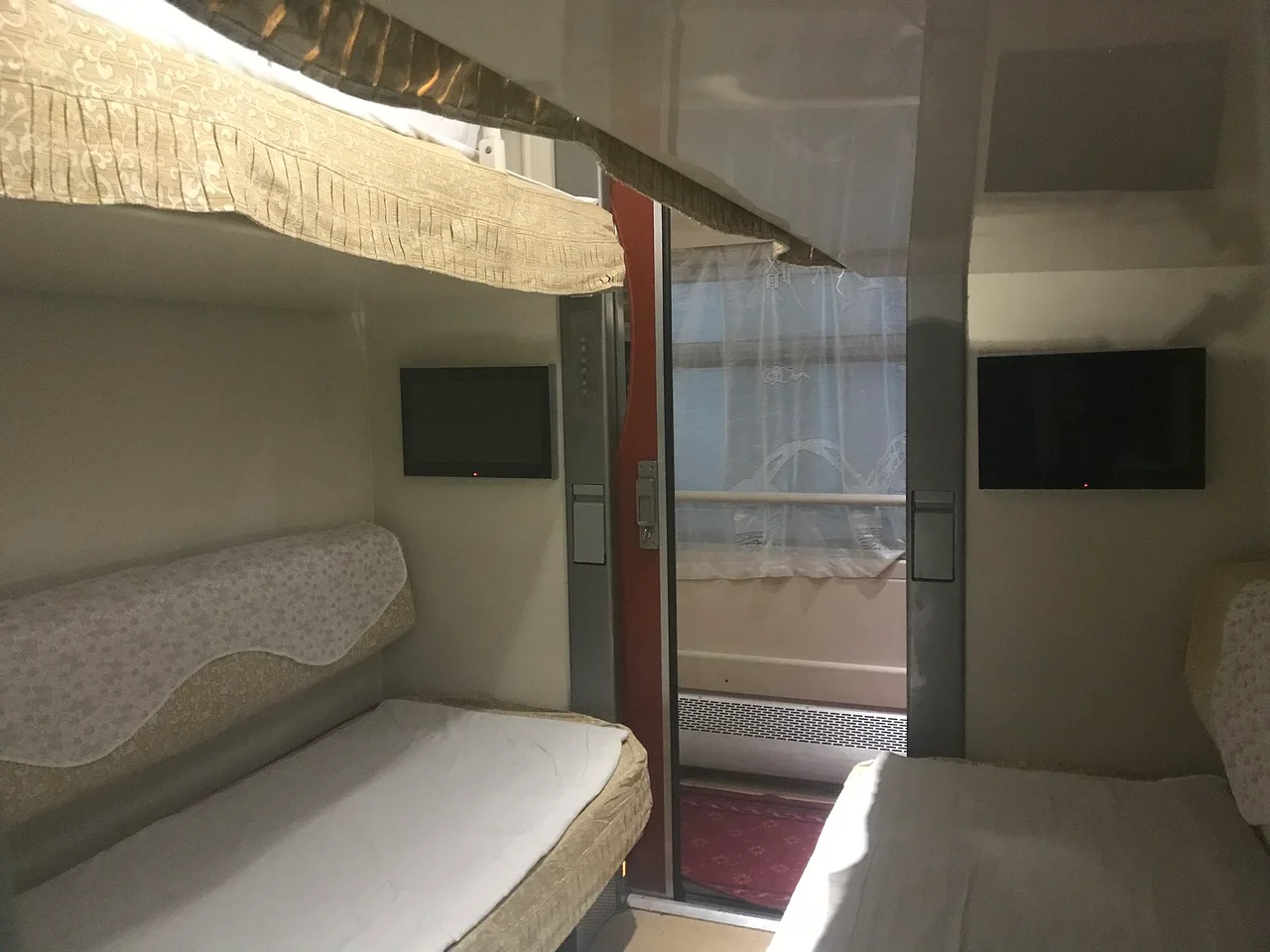 A soft-sleeper compartment with a lower bunk made up with a pale blanket, a small TV screen and a window