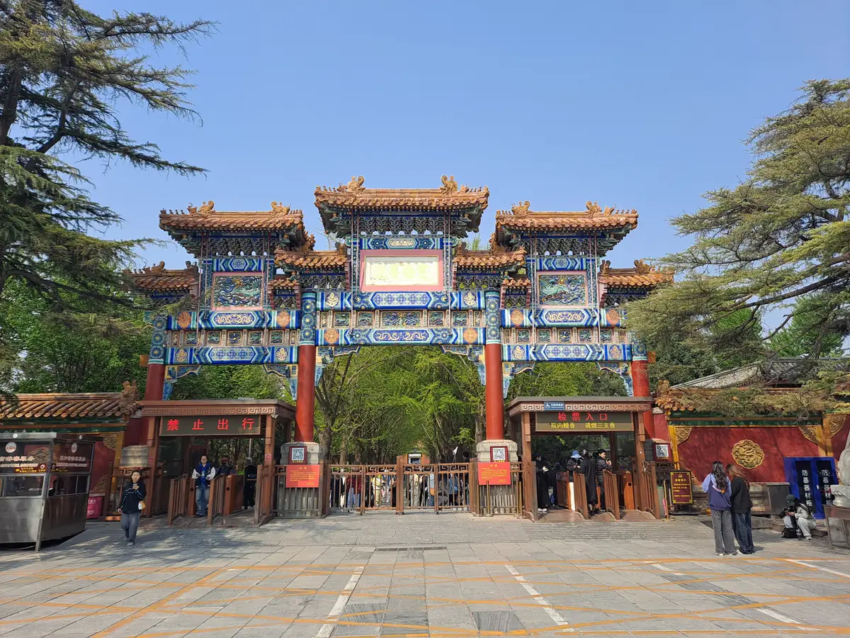 A painted wooden archway with three roofs at the entrance to the Lama Temple, framed by trees