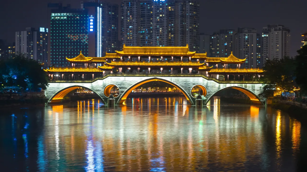 The covered Anshun Bridge lit gold at night, its arches and pavilion roofs reflected in the river, with lit tower blocks behind