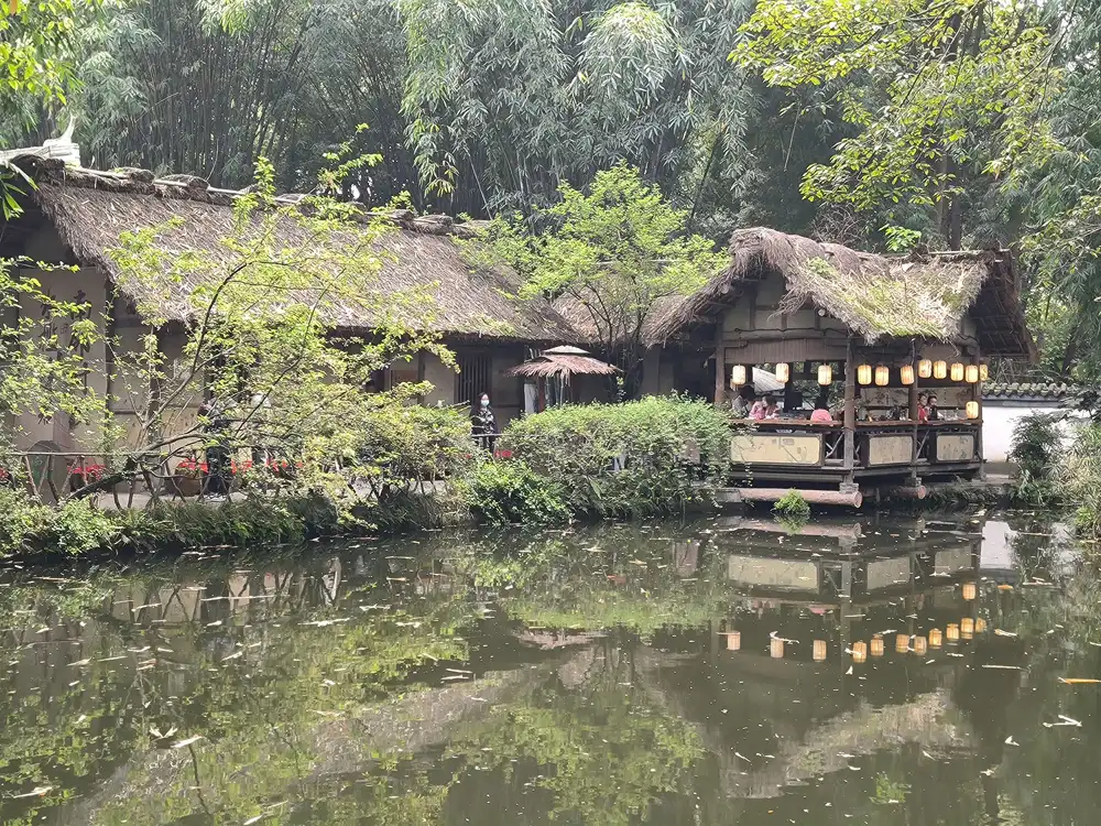 Low thatched-roof pavilions among dense trees and bamboo, reflected in a still green pond