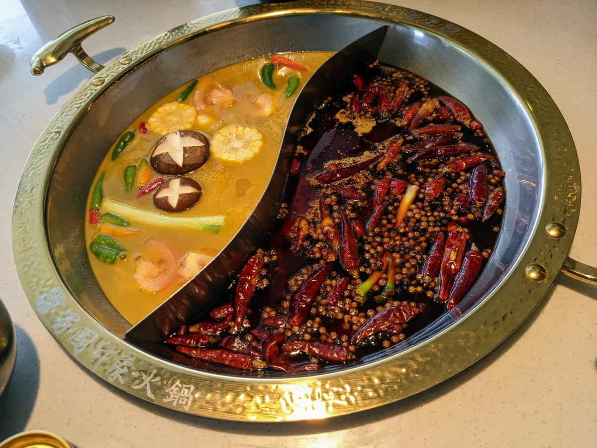 A round brass hotpot divided into two halves, one a mild broth with mushrooms and prawns, the other packed with dried red chillies and peppercorns