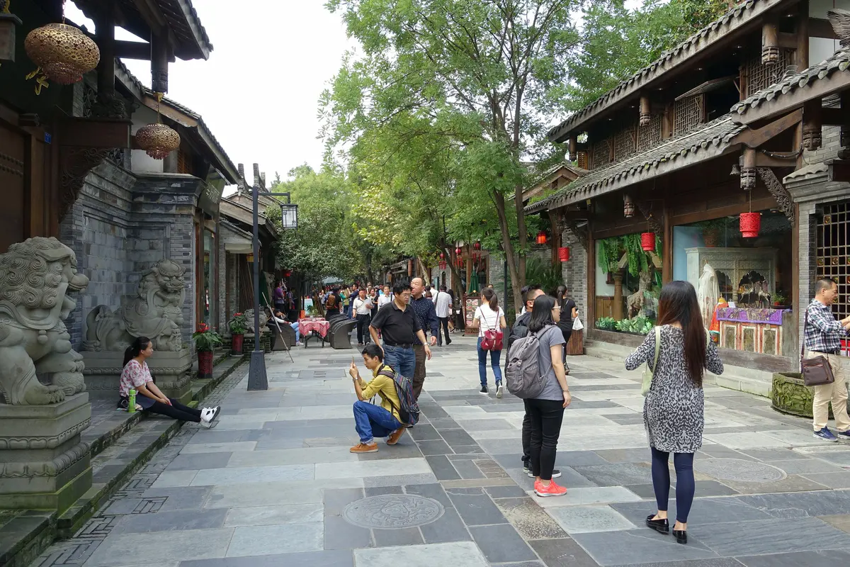 A paved pedestrian lane with stone lions, grey brick walls and wooden-fronted two-storey houses under trees, with visitors taking photos
