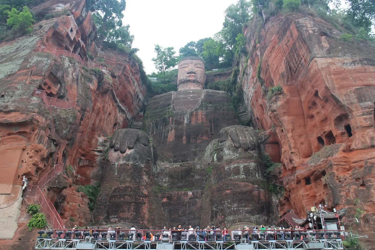 A giant seated Buddha carved into a red sandstone cliff, with a crowd of visitors on a platform at its feet for scale