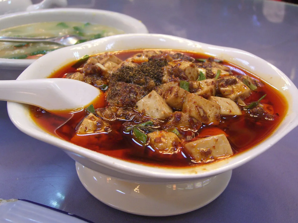 A white bowl of mapo tofu: soft tofu cubes and minced meat in a red chilli-oil sauce, dusted with ground pepper