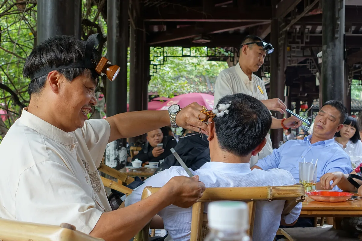 A man wearing a head torch cleans the ear of a seated customer at an outdoor teahouse table, with other tea drinkers around them