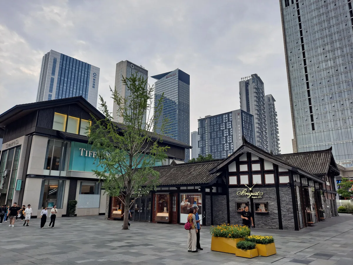 A paved square with low traditional Sichuan-style shop buildings in front of a cluster of glass office towers under a grey sky
