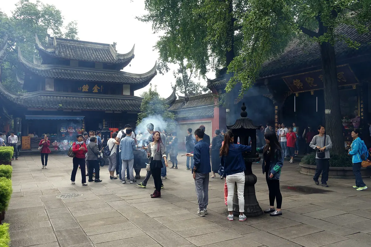 Visitors and worshippers in a paved temple courtyard with incense smoke rising from a burner in front of a multi-tiered pavilion