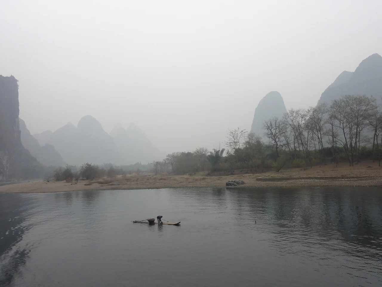 A fisherman on a small bamboo raft on the grey Li River with misty limestone hills behind