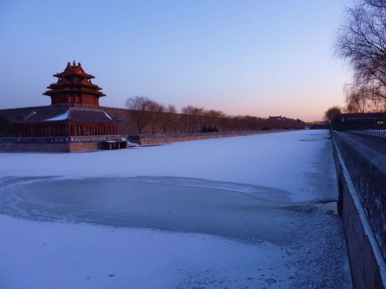 A red corner tower of the Forbidden City beside a frozen moat covered in snow under a violet dusk sky