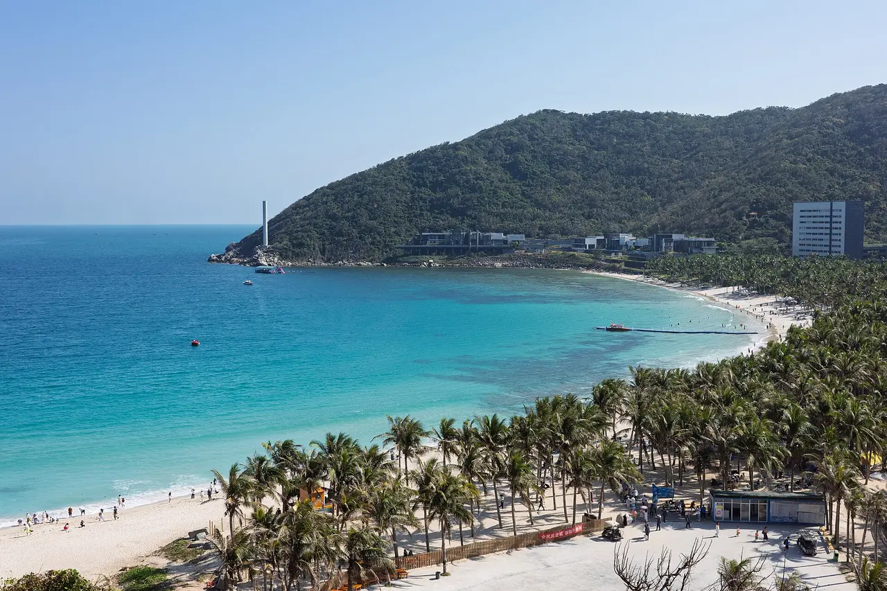A curve of white sand beach with turquoise water, palm trees and a green headland under a blue sky