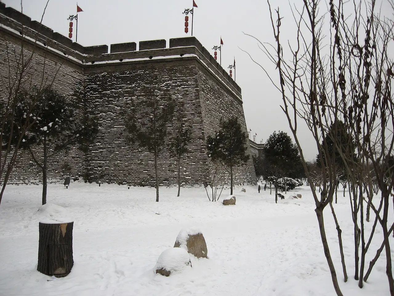 A dark brick city wall with battlements and watchtowers seen across a snowy park with bare trees