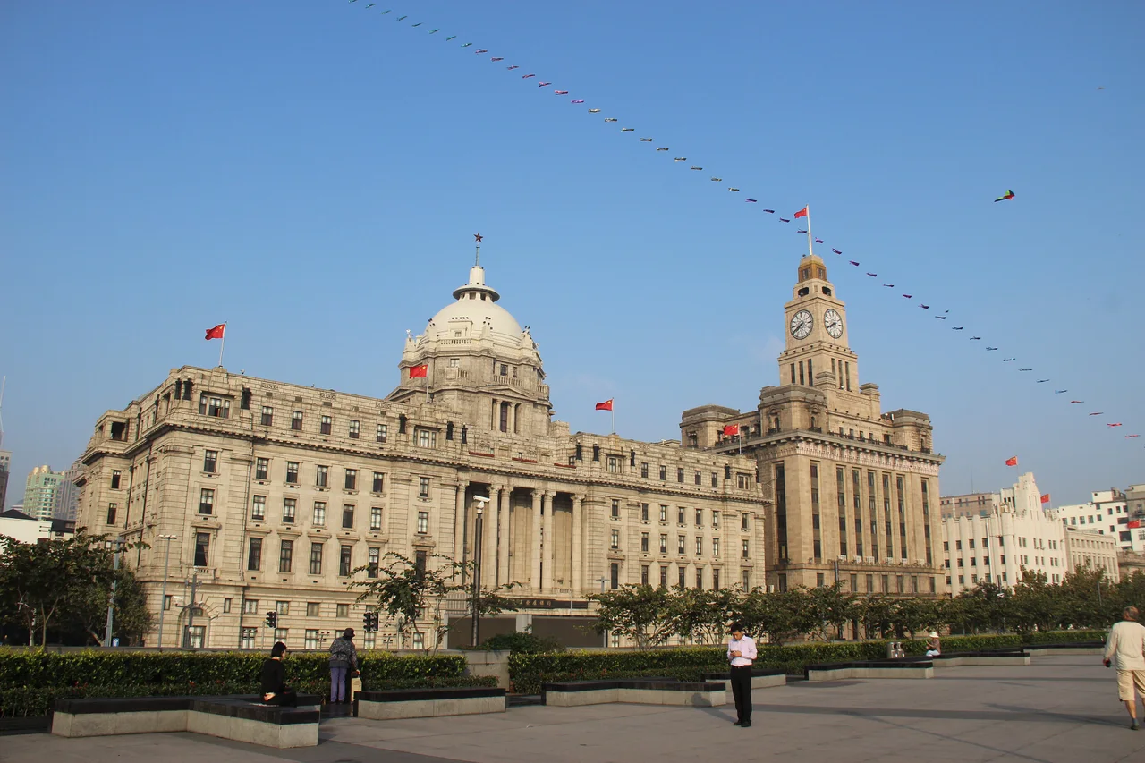 The domed former Hong Kong and Shanghai Bank building and the Customs House clock tower on the Bund, red flags flying, a row of kites strung across a blue sky