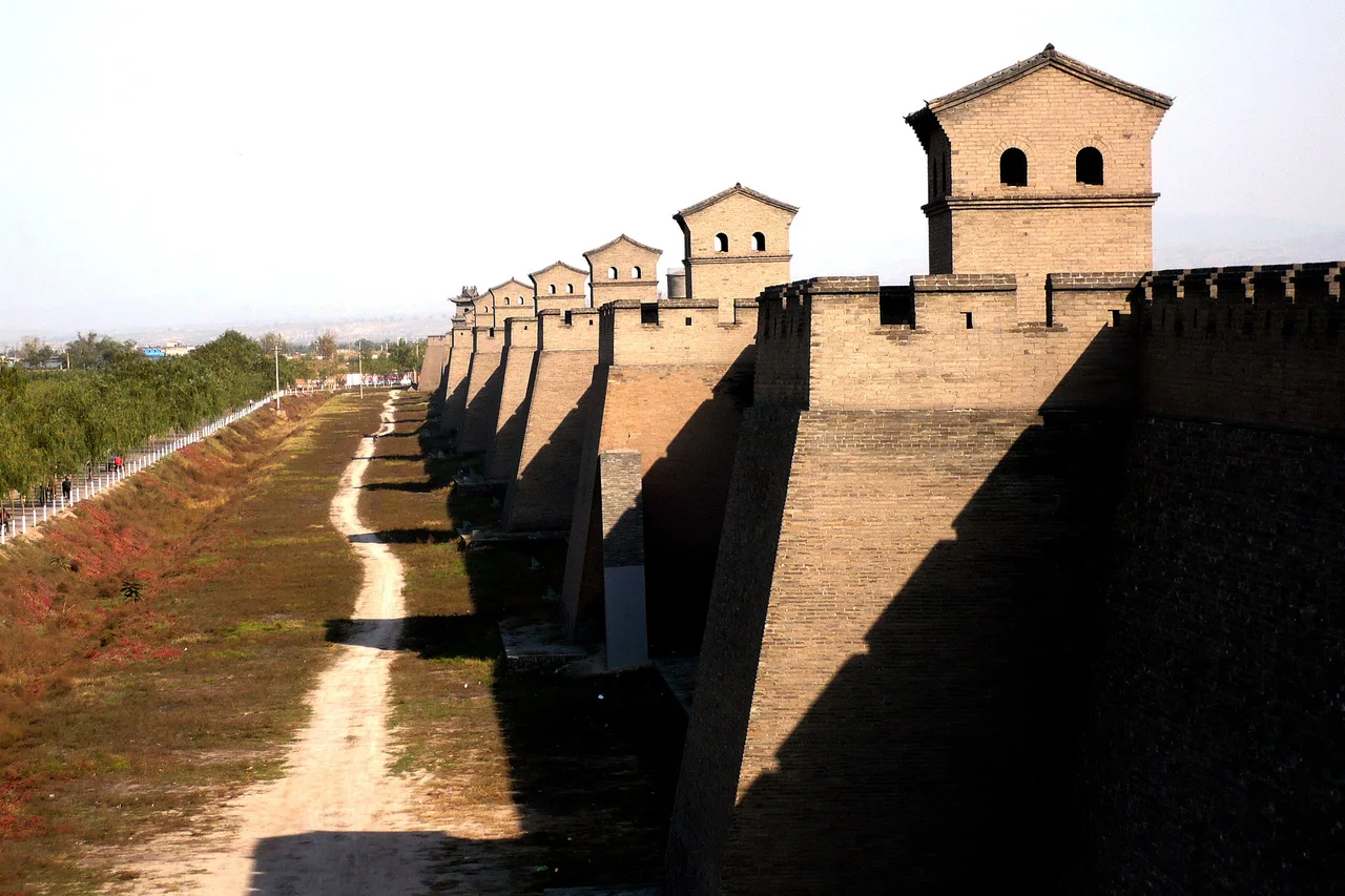 The outer face of Pingyao's grey brick city wall with a long row of square watchtowers, a dirt path and grass along the foot of the wall