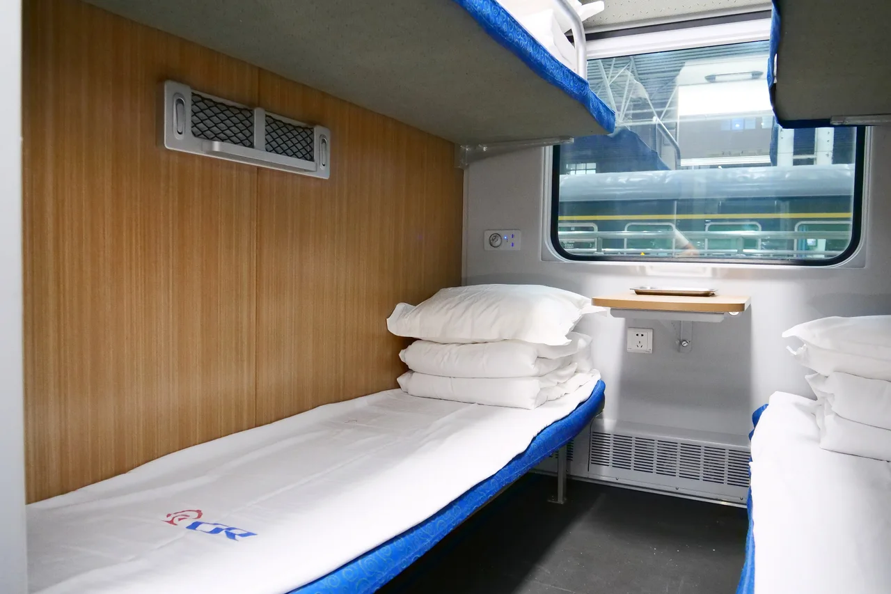 A made-up lower berth with white pillows and a folded duvet in a sleeper compartment, wood-panelled wall, a small table under the window and upper bunks above