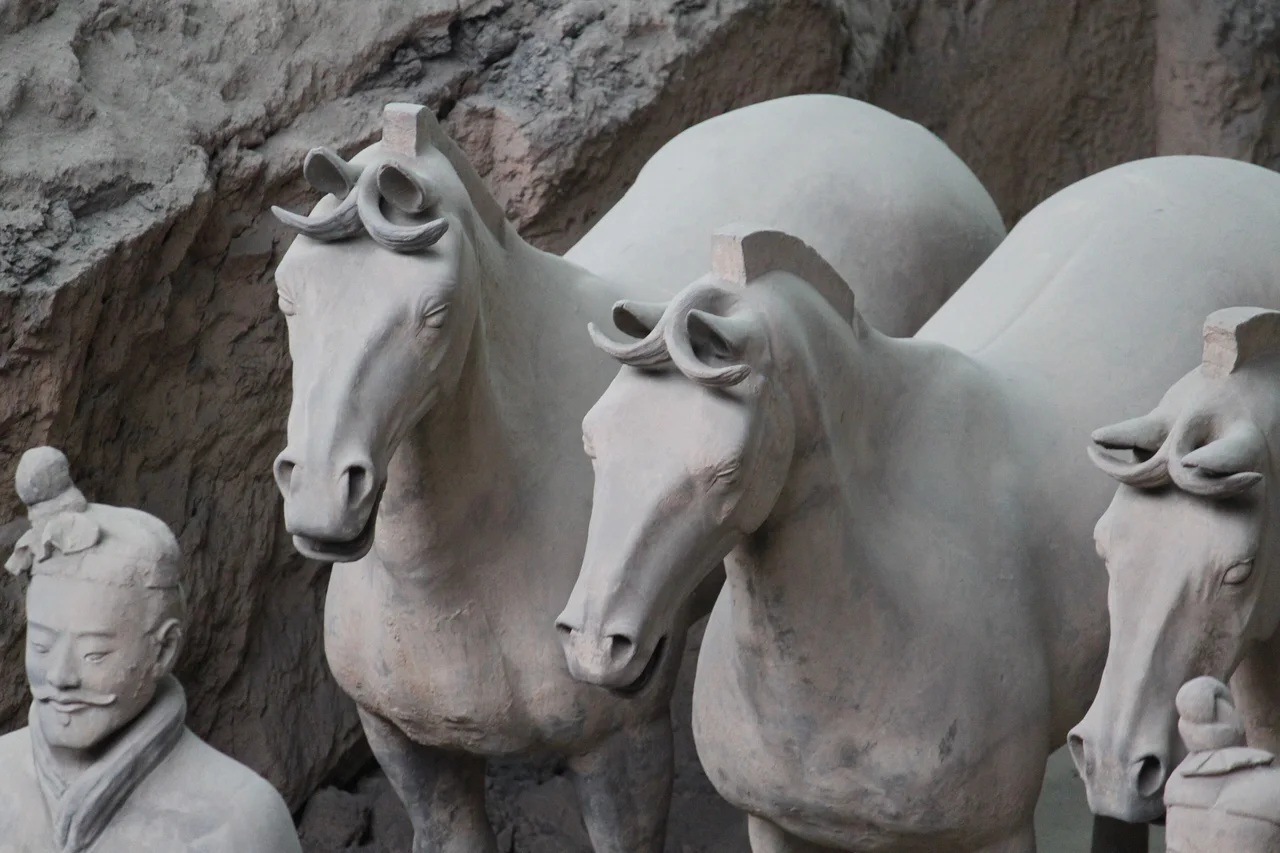 Life-size terracotta horses with their manes cut short, standing in a row beside a terracotta soldier in an earthen trench