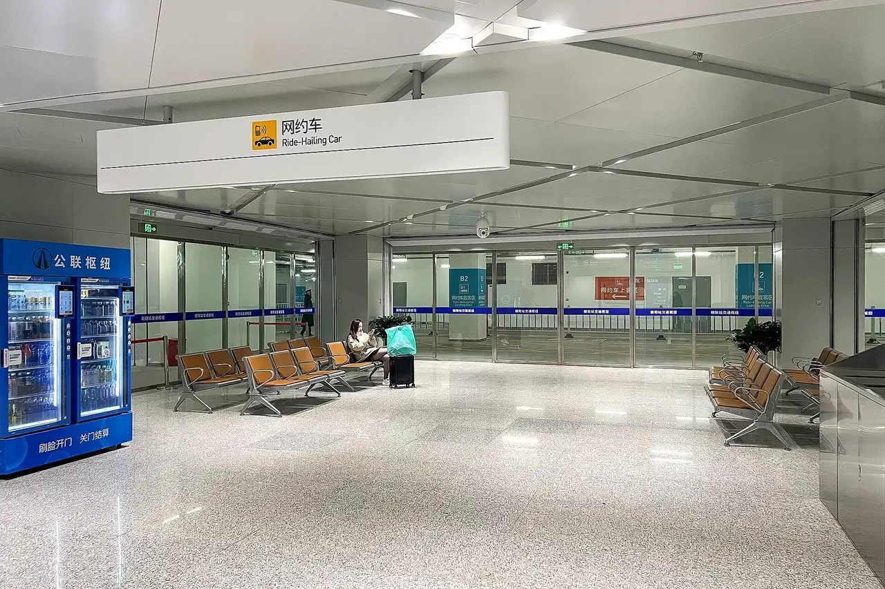 A bright underground hall at Beijing Chaoyang railway station with a sign reading Ride-Hailing Car above glass doors and rows of seats
