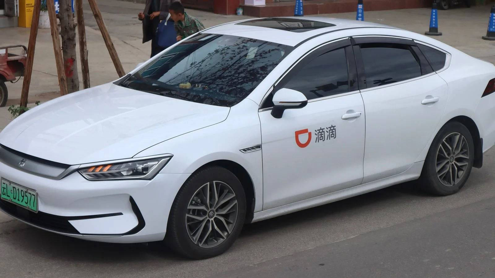 A white electric saloon car with the orange DiDi logo and the characters for Didi on its front door, parked at a kerb