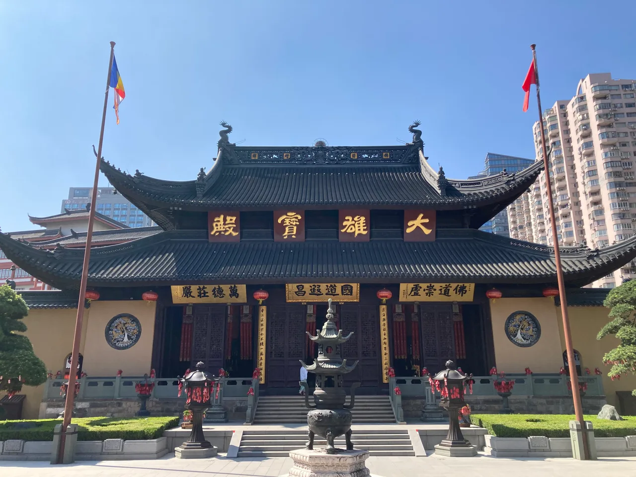 The main hall of Jade Buddha Temple in Shanghai with a flag pole, incense burners and high-rise buildings behind