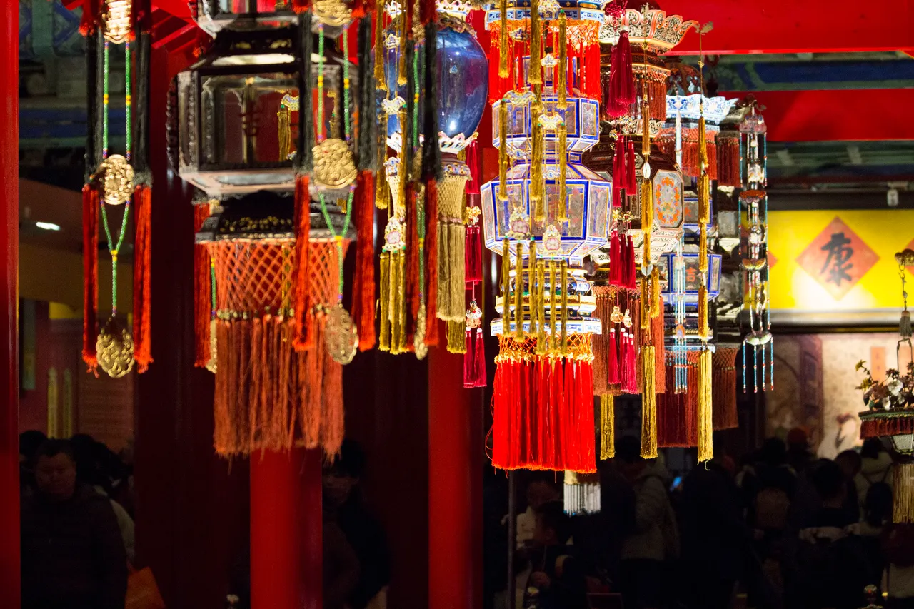 Hanging palace lanterns with red and gold tassels in a dim hall of the Forbidden City, visitors behind them