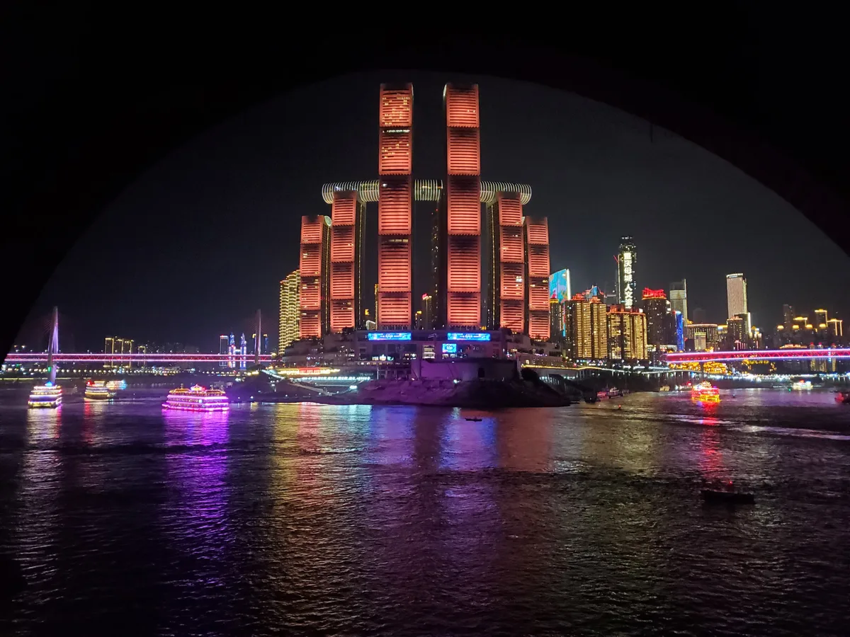 The lit Raffles City towers and a bridge reflected in the river at night, with brightly lit cruise boats on the water