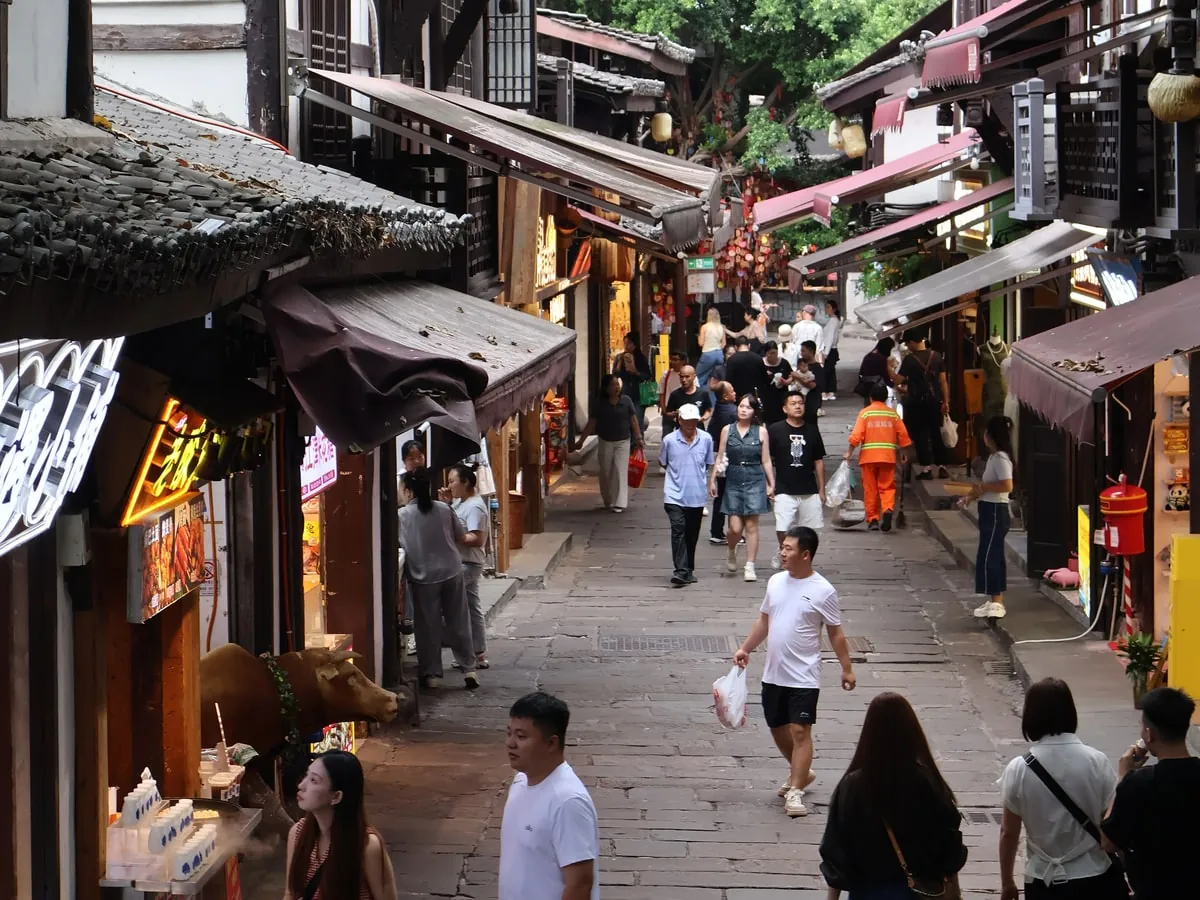 A stone-paved lane between dark timber shophouses with tiled eaves and maroon awnings, busy with people walking and browsing the stalls