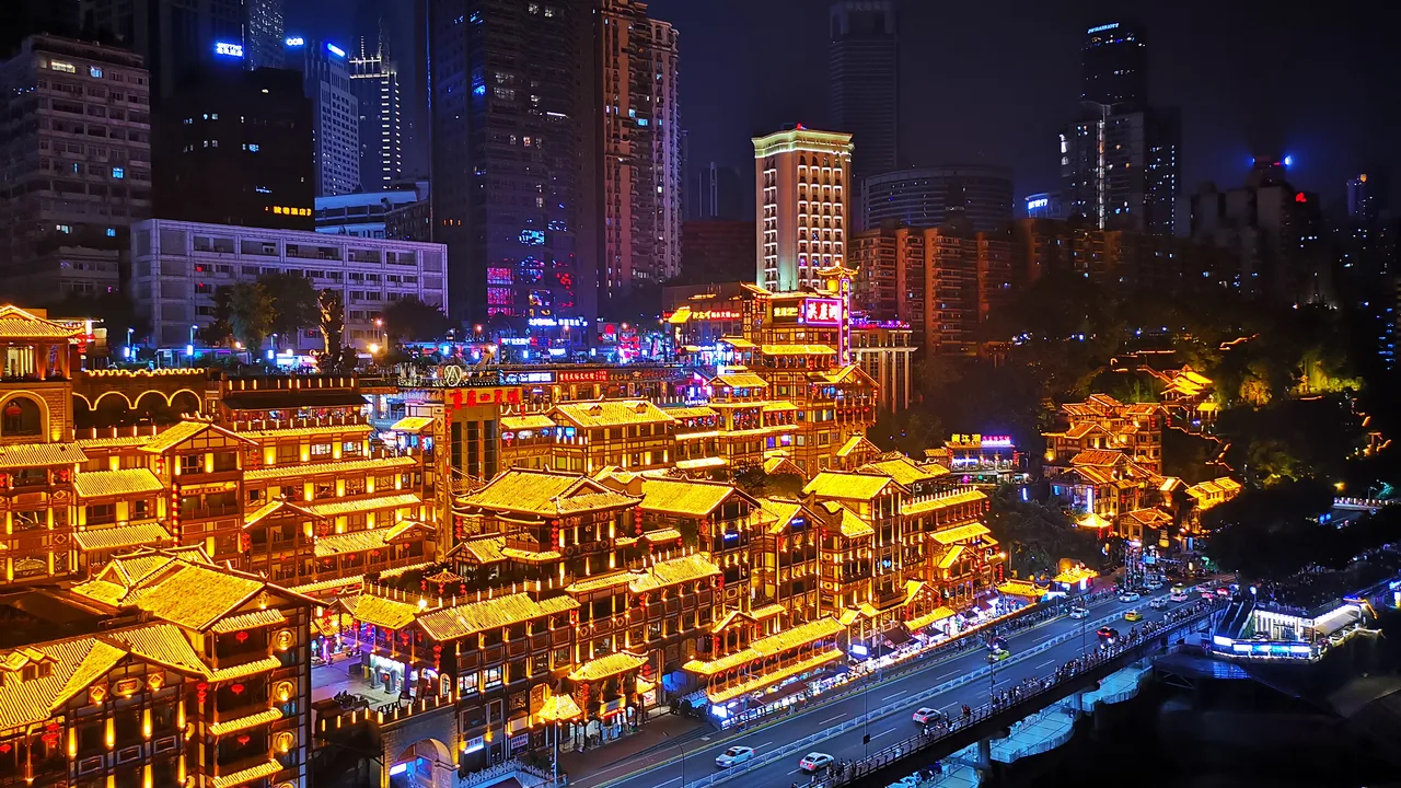 The stilt-house complex of Hongyadong lit gold at night, stepping down a cliff to a riverside road, with tower blocks behind