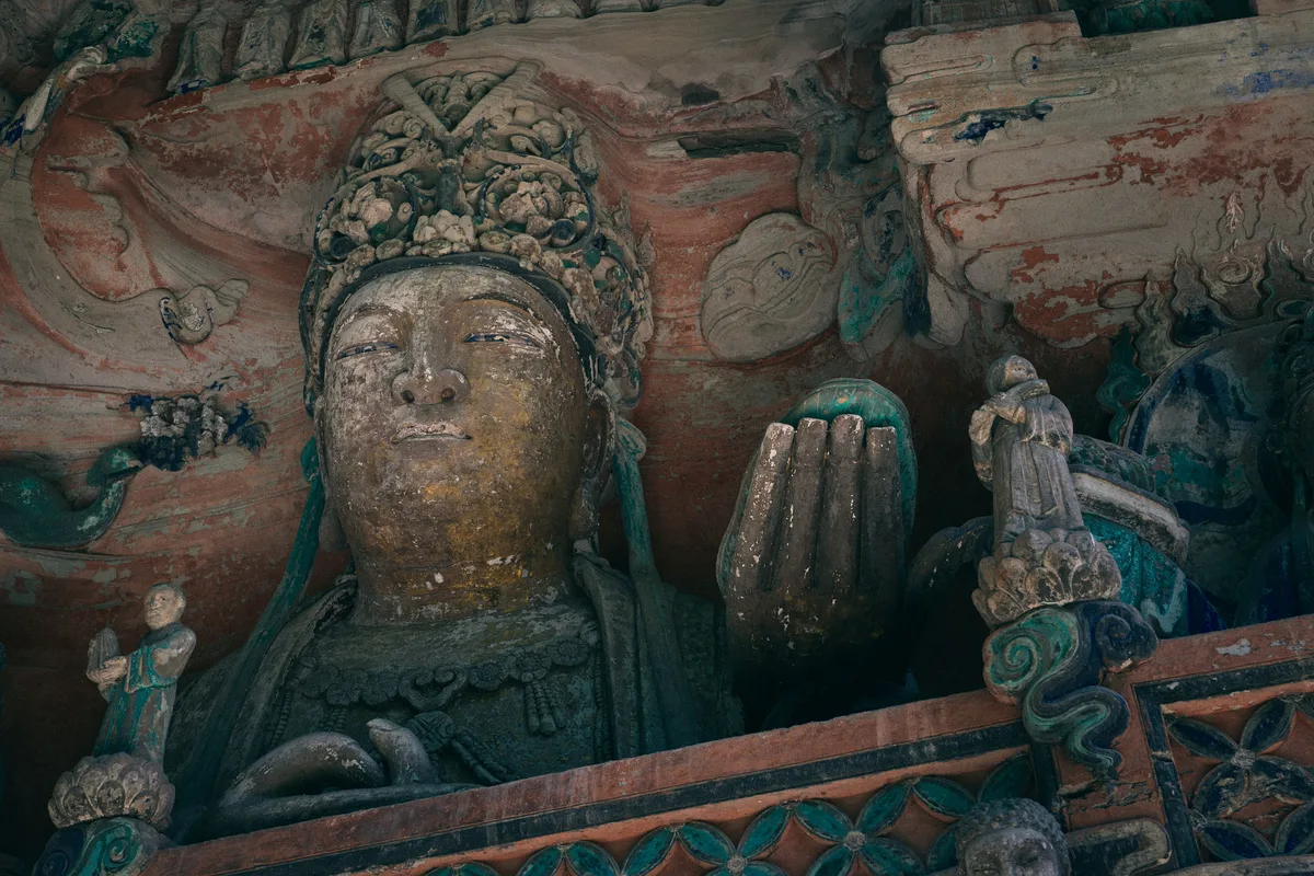 A carved and painted stone figure of a crowned bodhisattva with raised hands set into a rock face, traces of red and green paint around it