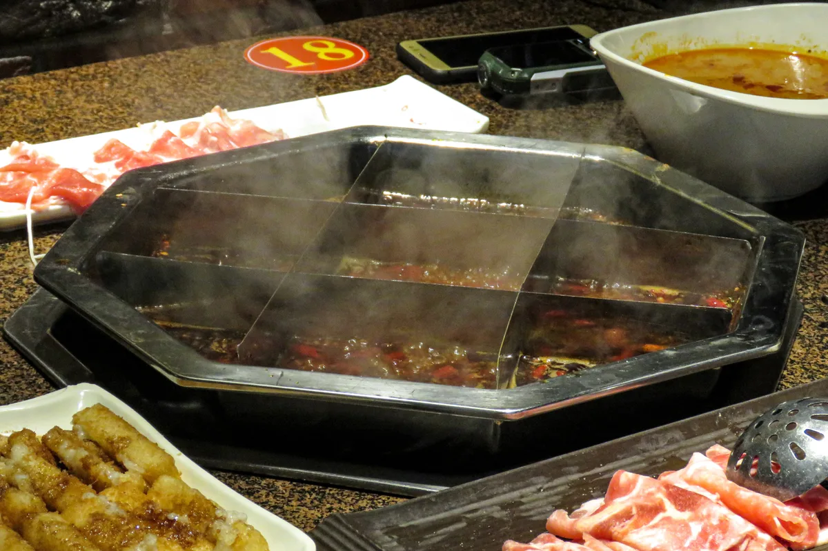 A steaming octagonal hotpot divided into a grid of compartments of red chilli broth, with plates of sliced meat around it