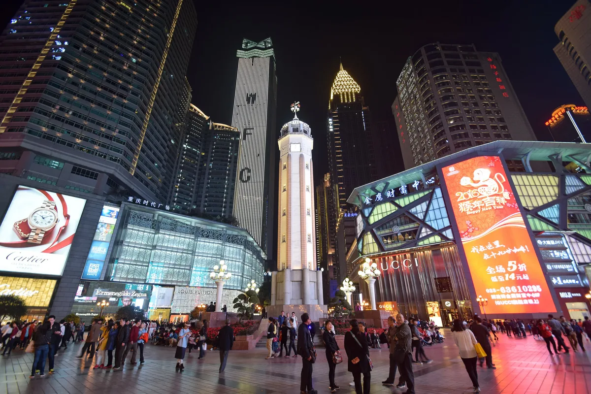 A white clock tower in a crowded pedestrian square at night, surrounded by skyscrapers and brightly lit shop fronts