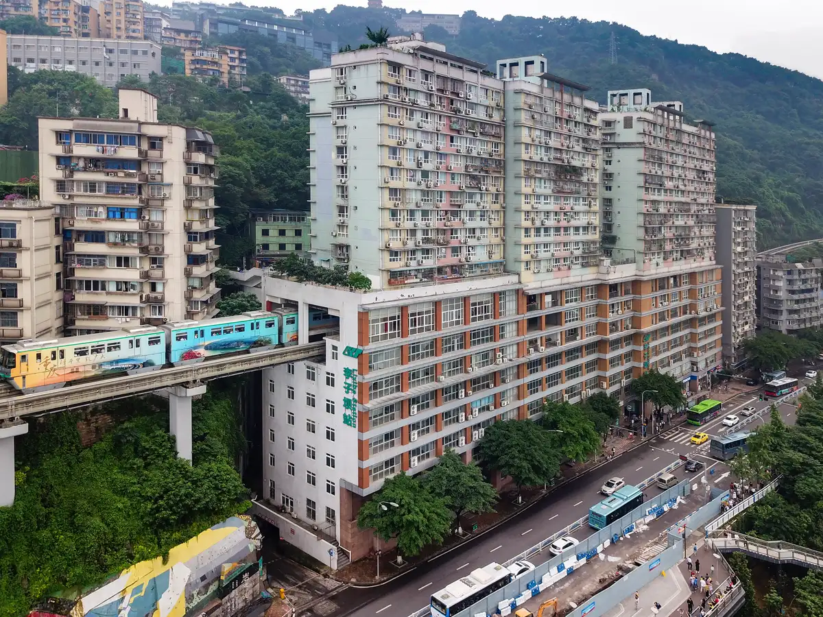 A light-blue monorail train entering a tall pale residential block at its lower floors, with traffic on the road beneath and hills behind