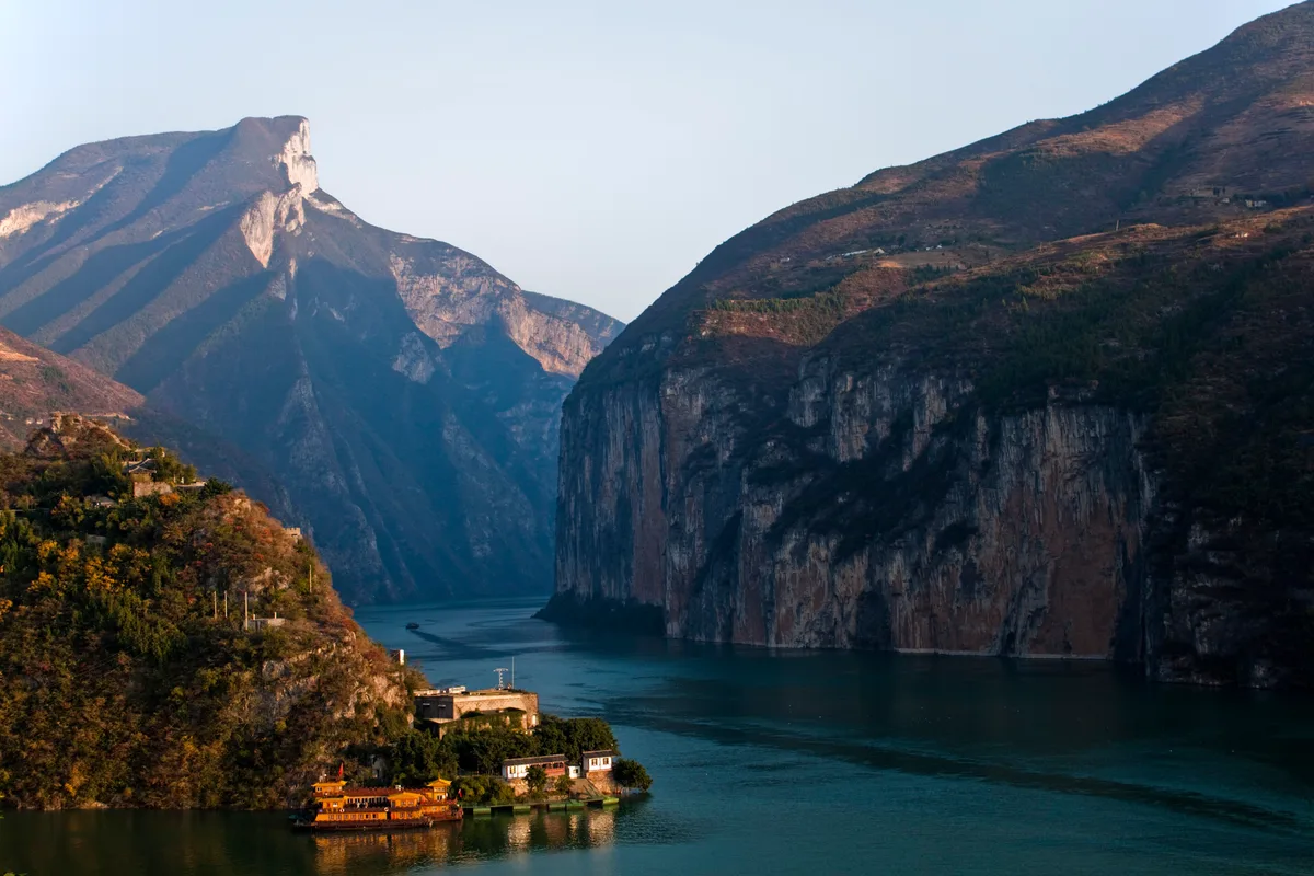 Steep cliffs of a narrow river gorge in late light, with temple buildings on a wooded spur above blue-green water