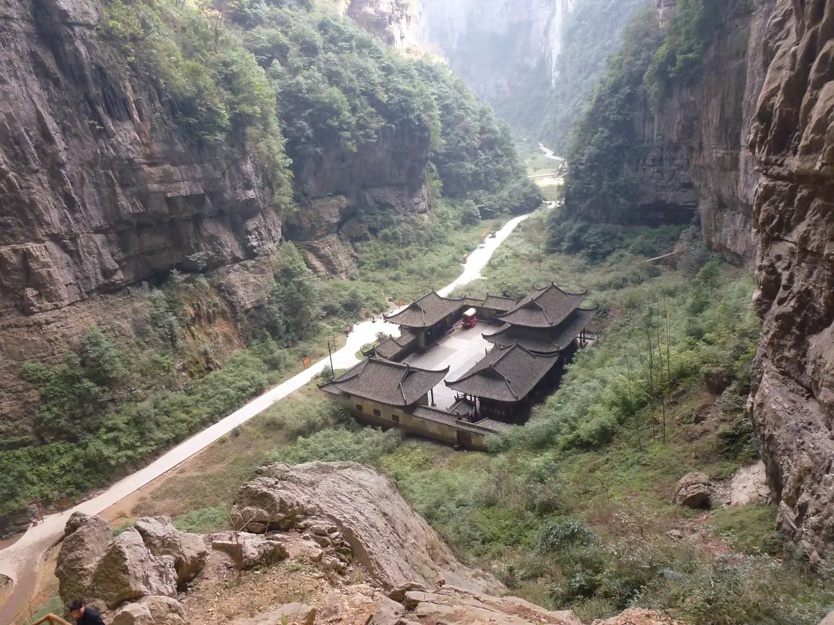 A deep gorge with sheer rock walls and a traditional inn with dark tiled roofs on the valley floor beside a footpath