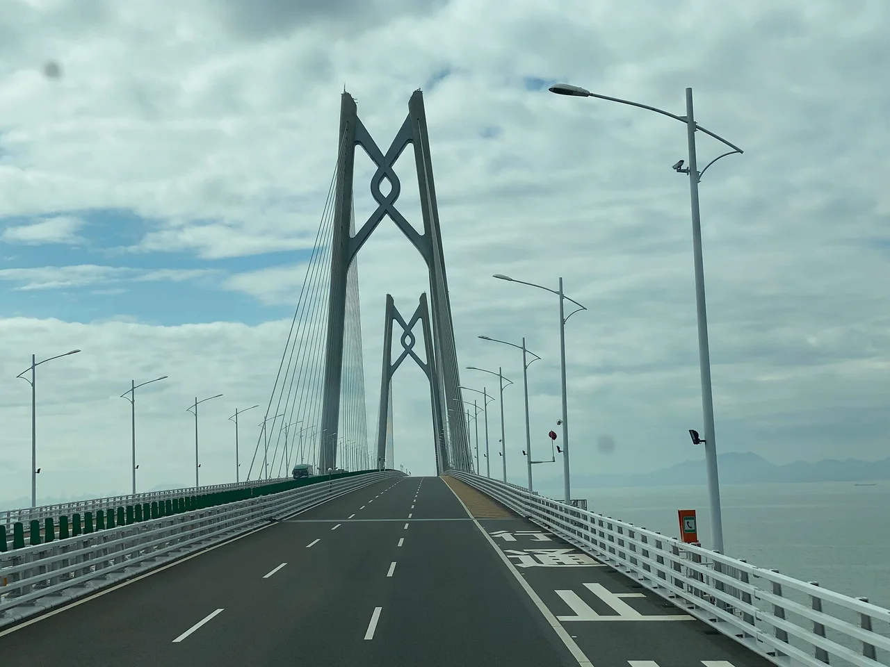 The tall cable-stayed tower of the Hong Kong-Zhuhai-Macao Bridge seen from the roadway, with lamp posts and an overcast sky