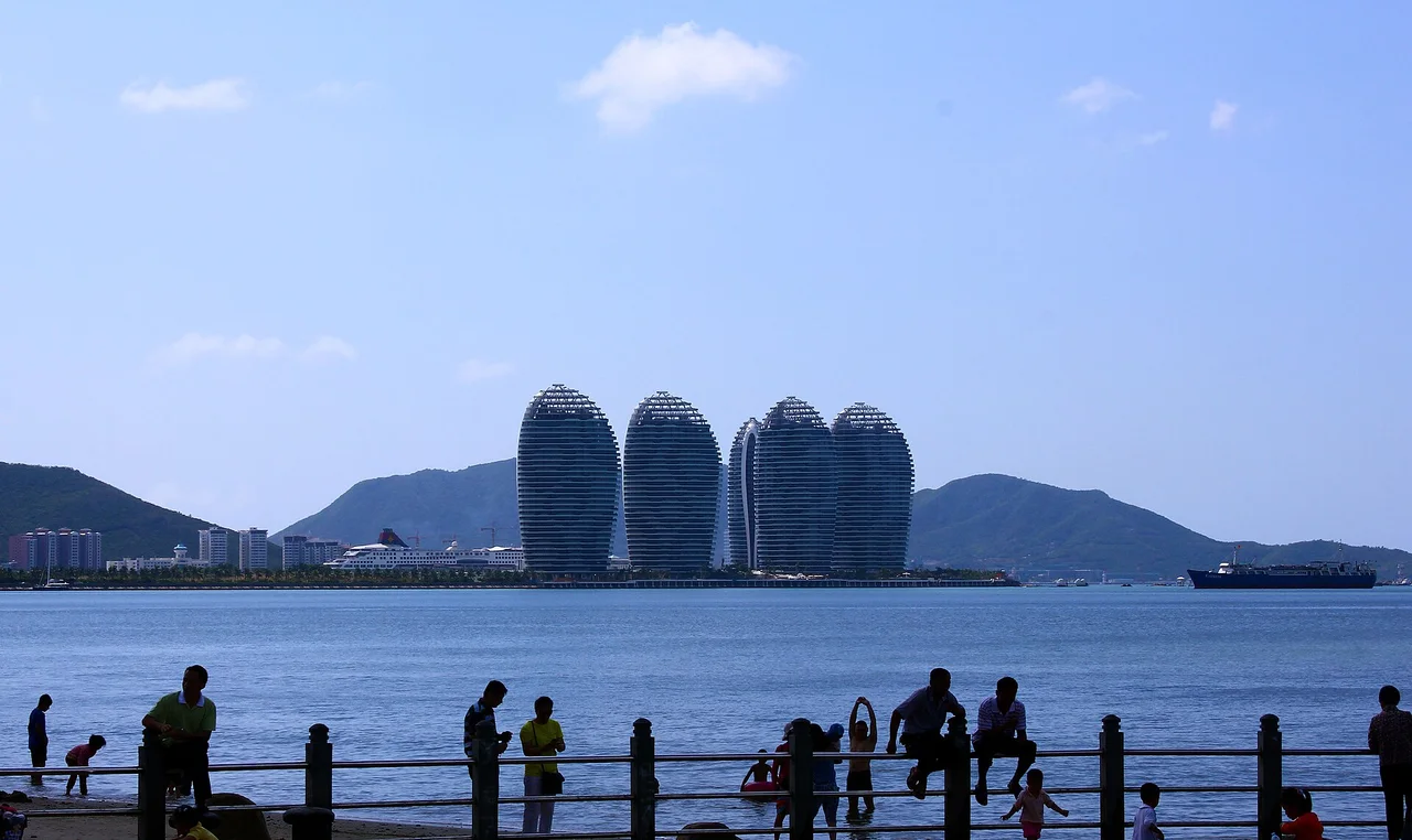 The tall rounded towers of Phoenix Island across the water from Sanya, with people standing along a seafront railing