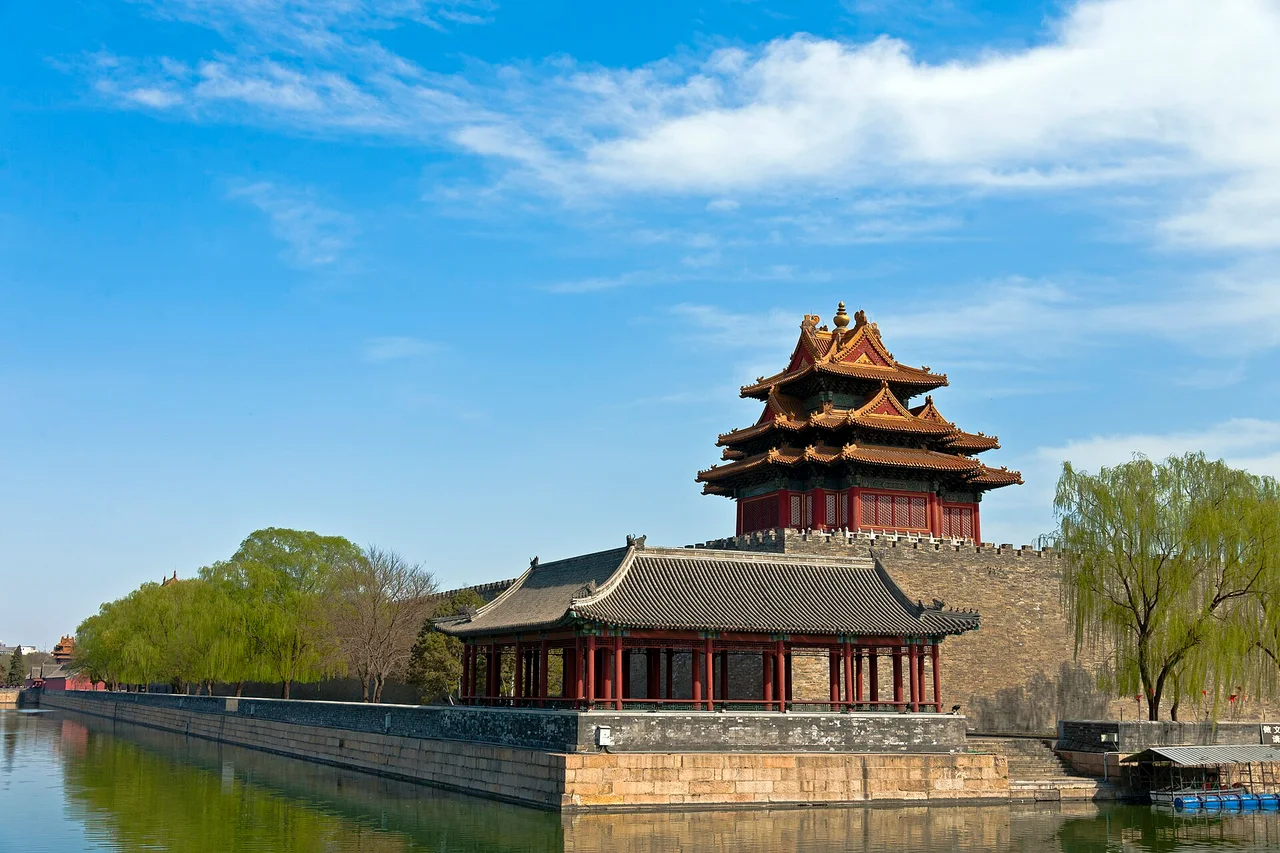 The northwest corner tower of the Forbidden City above the palace wall, reflected in the moat, with willows along the bank