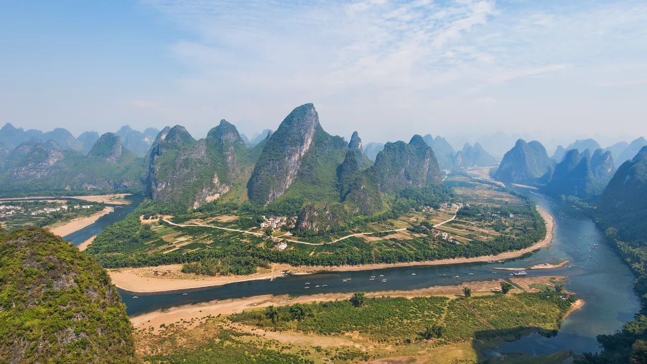 Green karst peaks rising above a wide bend of the Li River, with sandbanks, small boats and a farming village on the flat ground between the hills