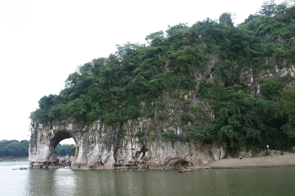 A rocky hill covered in trees with a natural arch at its base dipping into the river, like an elephant drinking