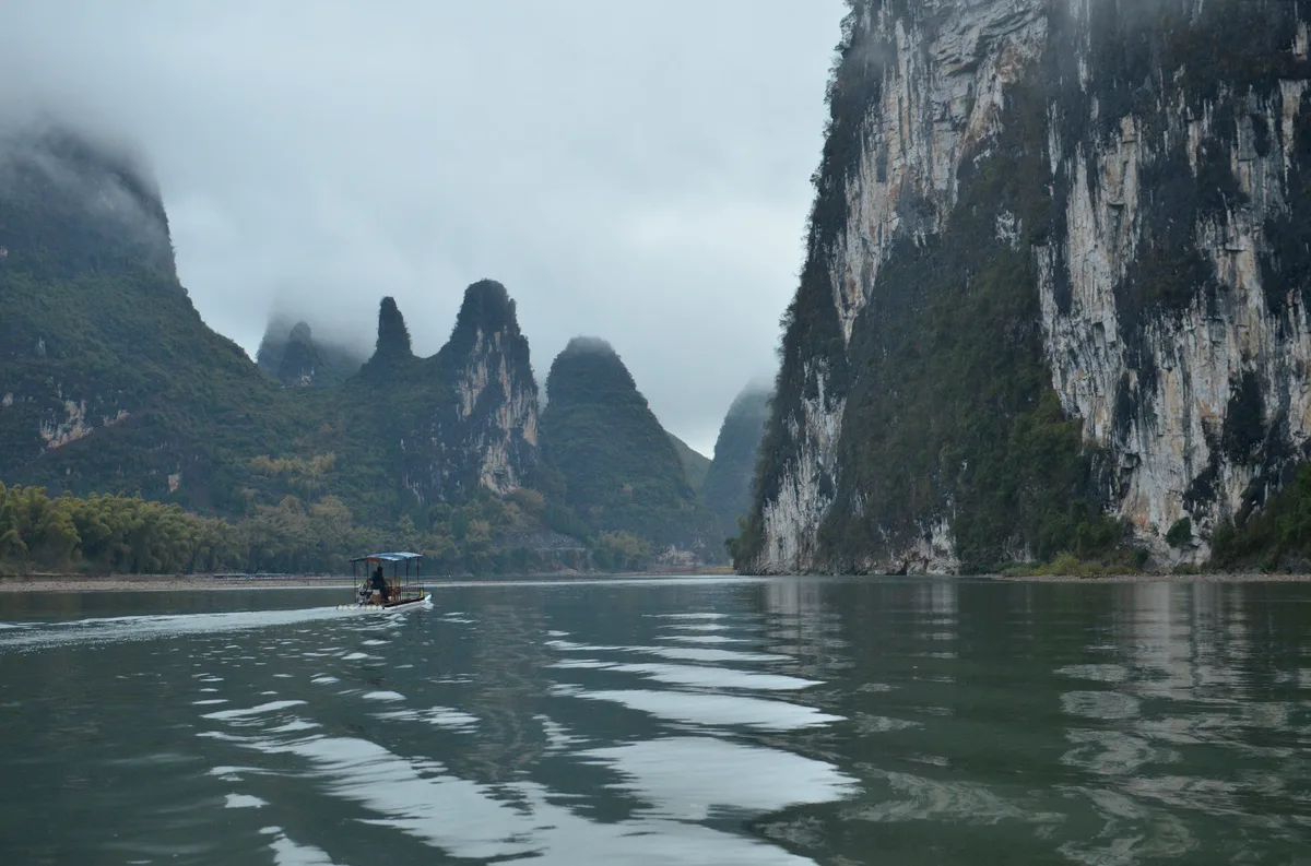 Sheer grey limestone cliffs dropping into a calm green river under low cloud, with a small boat crossing