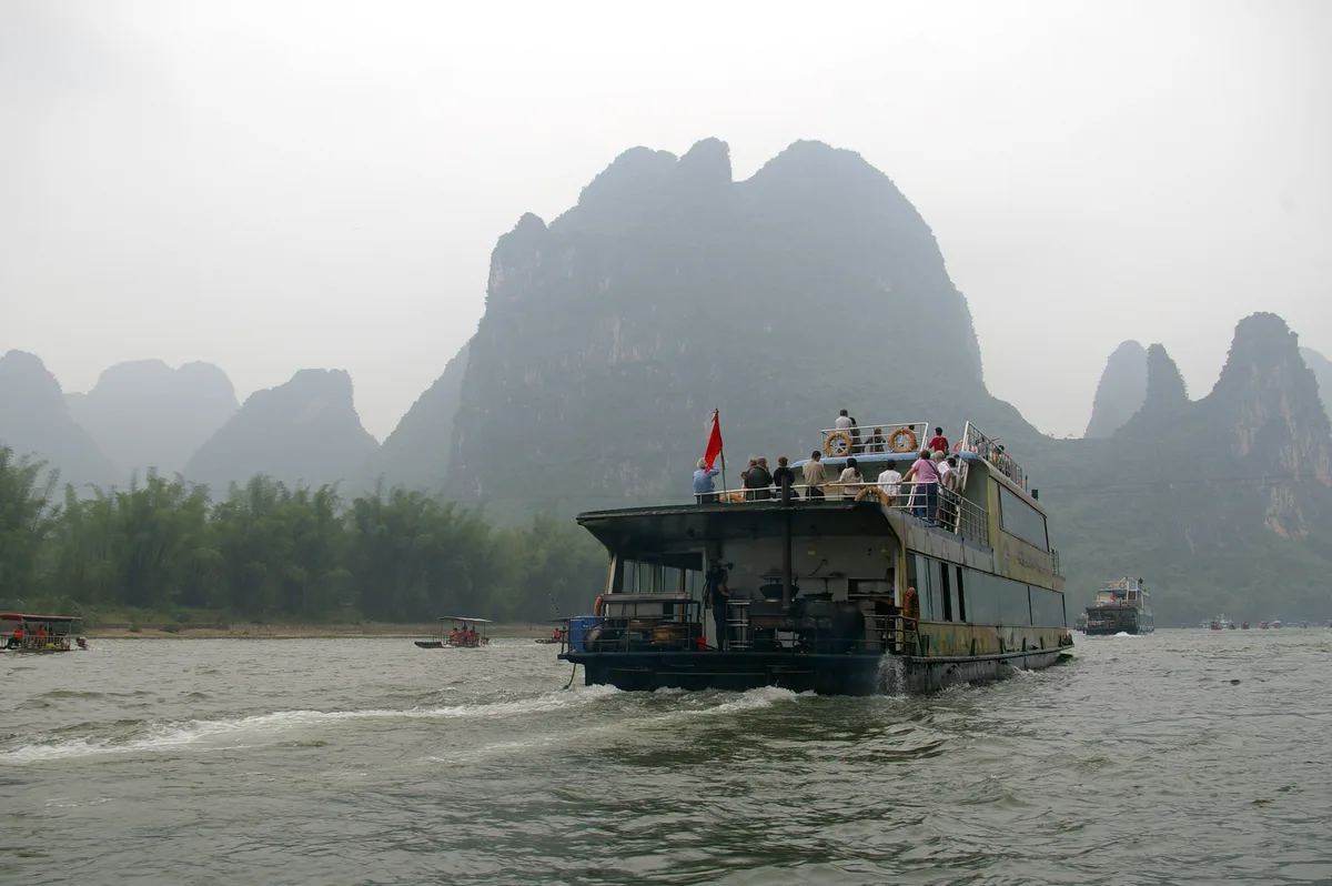 A white two-deck cruise boat with passengers on its top deck sailing down a wide river towards misty karst peaks