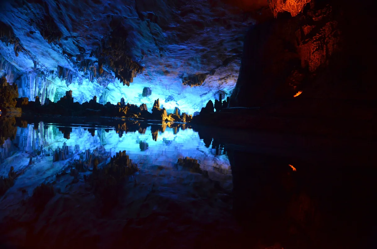 Rock formations lit blue and red inside a cave, reflected in a still pool
