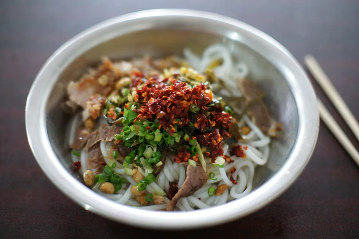 A metal bowl of white rice noodles topped with sliced meat, peanuts, chopped spring onion and chilli flakes, with chopsticks beside it
