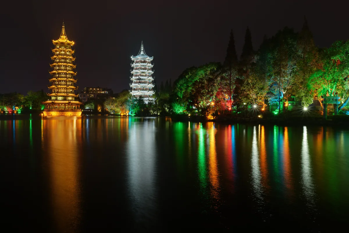 Two tall pagodas, one lit gold and one silver, reflected in a lake at night, with coloured lights in the trees beside them