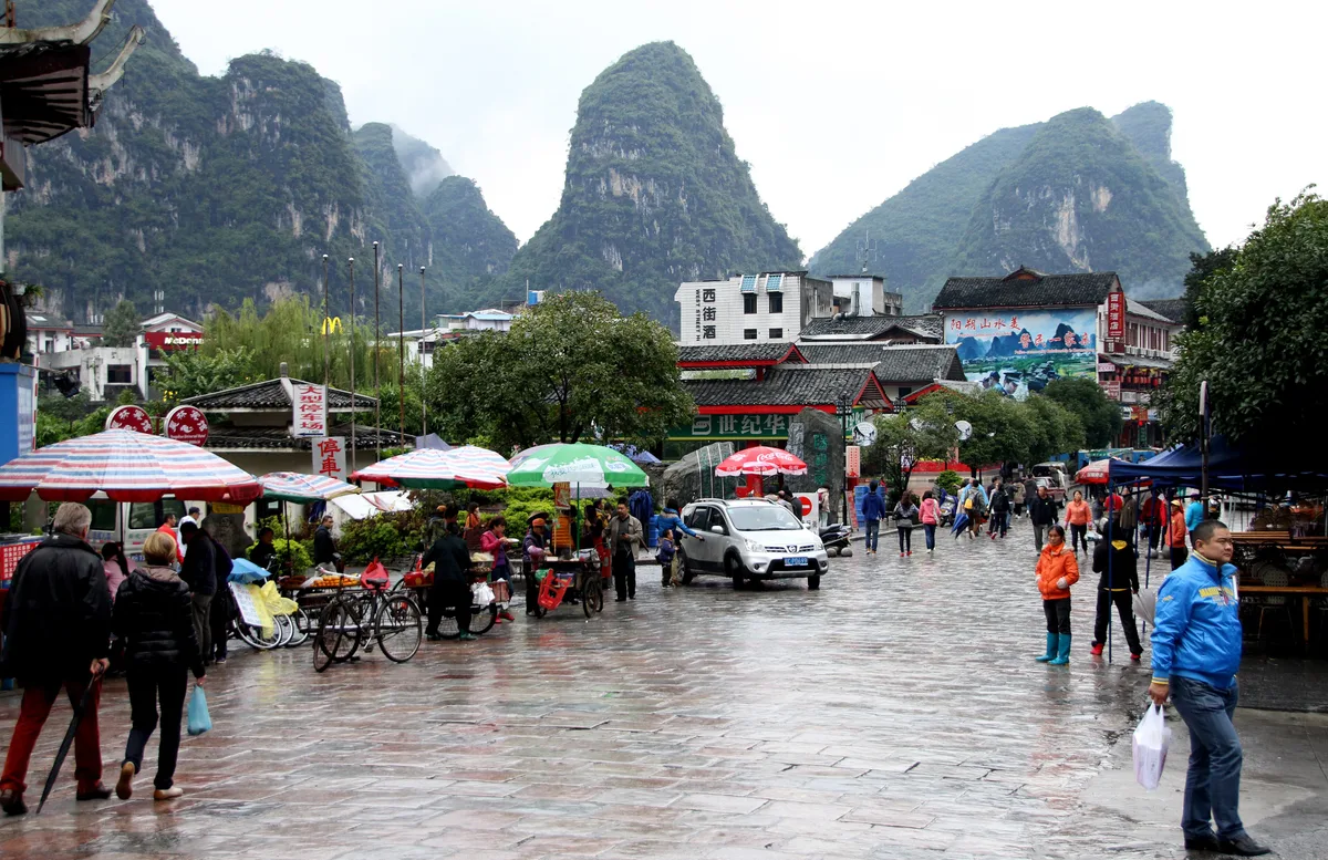 A wet stone-paved street with shop fronts and umbrellas, and tall karst peaks rising directly behind the buildings