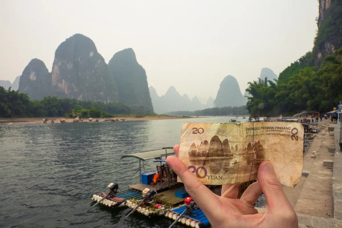 A hand holding up a 20-yuan banknote in front of the matching view of karst peaks across the river, with a moored raft below