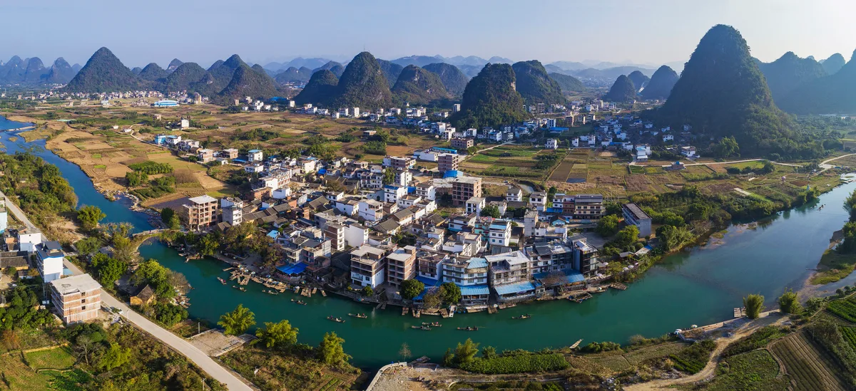 Aerial view of a village of white houses on a river bend, surrounded by green fields and a ring of karst peaks
