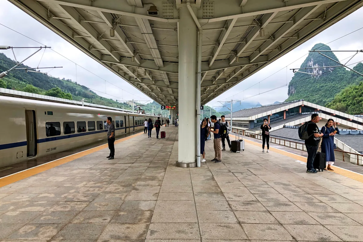 A long covered platform with a white high-speed train on one side and a karst peak rising behind the station roof