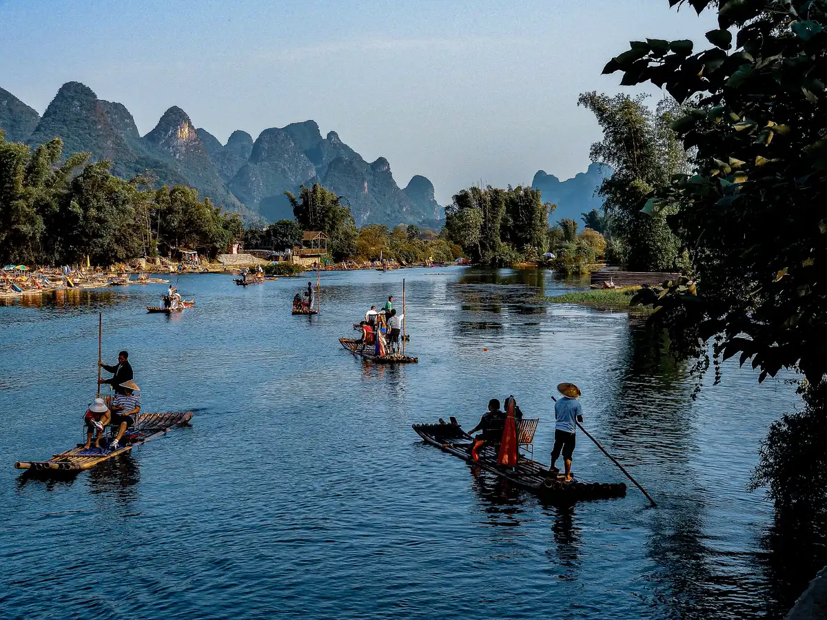 Several bamboo rafts with boatmen and passengers drifting on a calm blue river, with karst peaks on the horizon