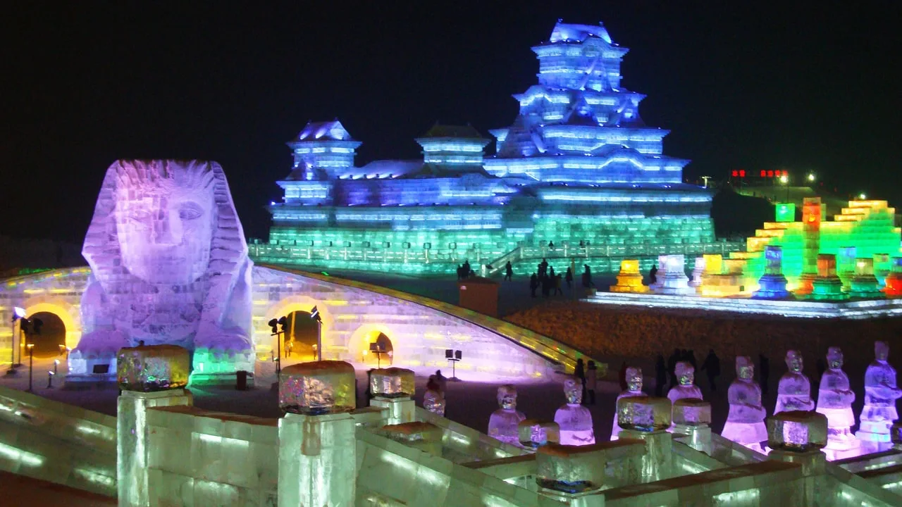 A lit ice sphinx and an ice castle with a pagoda-style tower glowing blue and green at night, with rows of ice soldier figures in the foreground